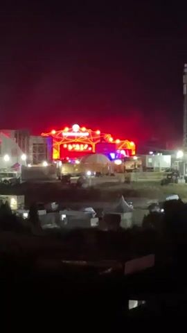The video shows a nighttime aerial view of an outdoor event venue, likely a music festival, with a large, brightly lit stage at its center. The stage is adorned with red and blue lights, creating a vibrant atmosphere. Surrounding the stage are various structures, including tents and other buildings, also illuminated by lights. Some vehicles are visible in the background. The overall scene suggests a lively event is underway or about to begin.