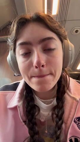 A young woman applies eyebrow gel while seated on a train. She faces the camera directly and wears headphones. Her hair is styled in pigtails. She has a nose ring and wears a necklace. She wears a pink jacket. A sign for "Priority Seats" is visible in the background.