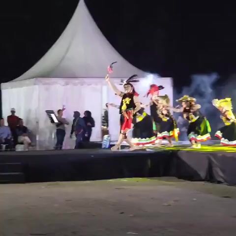 This is a video of a cultural performance taking place on a stage at night. A group of dancers, mostly women, are performing a traditional dance. They are wearing colorful costumes with skirts and headwear. One dancer in the center is wearing a black outfit with a headdress, and the others are wearing similar black dresses with red and green accents and straw hats. They are all performing synchronized movements, with some holding props like sticks. In the background, there is a white tent and a few people sitting in the audience on the left side. The stage is lit with yellow and white lights, and there is some smoke or fog effect creating a dramatic atmosphere. The overall scene suggests an outdoor cultural event.