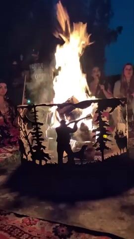 The video shows a bonfire in the center of the frame. The fire is large and bright, with flames reaching high into the air. Around the fire, several people are gathered. To the left, a woman in a colorful dress is looking towards the camera. To the right, two women are sitting and appear to be talking or singing. In the background, a person wearing a t-shirt with text on it is visible. The bonfire is contained within a metal fire pit that has a cut-out design depicting a forest scene with a hunter and a deer. The ground around the fire pit is covered with patterned rugs. The overall setting appears to be outdoors at night, with trees visible in the background and a dark sky above.