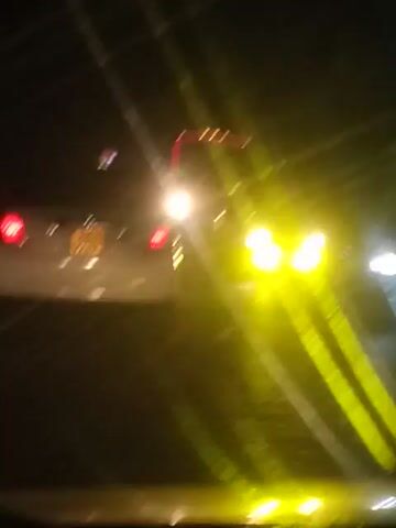The video appears to show a car crashed on its side at night. The scene is dark, lit by overhead lights and the car's tail lights. The car is white and the license plate is visible. The video is shaky and the focus is unclear.