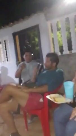 The video shows a group of people sitting outdoors at night. Several are seated on red plastic chairs.  They appear to be engaged in conversation.  One person is holding a plate with food.  The location appears to be a courtyard or patio, with buildings visible in the background. The lighting is dim, and some of the details are blurry.