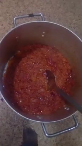The video shows an overhead view of a pot filled with a reddish-orange mixture, possibly a sauce or stew. A metal spoon is resting in the pot, partially submerged in the mixture. The mixture appears to be chunky and contains visible pieces of what could be onions or tomatoes. The pot has two metal handles and is sitting on a surface that looks like a countertop or table. Towards the bottom left of the frame, a person's leg and foot are visible, wearing dark blue pants. The lighting in the scene is dim, suggesting it might be indoors. The video is shot from a high angle and is slightly blurry.