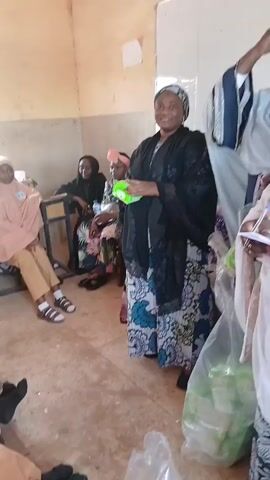 The media file captures a gathering of people, primarily women and girls, in what appears to be a classroom or community hall in Unguwar Shanu, Nigeria. The lighting suggests it's daytime, possibly mid-morning or afternoon, with natural light coming from a window. The room has plain, light-colored walls, and some tables and chairs are visible.

The central figures are several women, some of whom are dressed in traditional Islamic attire, including hijabs and abayas. One woman in the foreground, wearing a black outer garment over a patterned dress and a patterned headscarf, is holding a green object. She is smiling and appears to be addressing the group or presenting something. To her right, another woman in a light blue hijab and a white outer garment is also present, holding papers. In the background, several girls, likely students, are seated at desks, most wearing light brown hijabs and matching attire, looking towards the front. Their expressions are varied, some attentive, some perhaps a bit bored.

The activity appears to be some form of educational or community event. The woman in black might be a teacher or presenter, and the green object could be a gift or teaching aid. The presence of a whiteboard in the background further supports the idea of an educational setting. The overall atmosphere seems relatively calm and focused. The floor is a simple concrete material, and some debris is visible, suggesting a practical, perhaps under-resourced, environment. The attire of the individuals, particularly the hijabs and modest clothing, points to a community with strong Islamic traditions. No discernible text is visible in the media file.