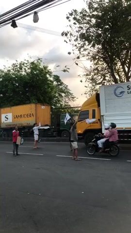 A group of people stand on a street next to two large trucks. One truck is yellow and has "SAMUDJERA" written on its side. The other truck is white and blue and has "Smart Logistics" written on its side. A motorcycle with two people on it drives past. One person in the group is holding a flag with a green cross on it. The other people are standing around and watching the trucks and the motorcycle. The sky is cloudy.