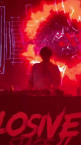 A DJ stands behind a console on a stage, illuminated by vibrant red lights and a large screen displaying abstract patterns and shapes, possibly flames or explosions. The stage has a banner across the front that reads "EXPLOSIVE STAGE" in a stylized font. There are bottles of water on the console. The overall atmosphere suggests a live music event or festival.