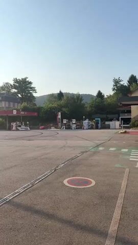This video shows an exterior view of a gas station and an adjacent building with large glass windows. The gas station appears to be empty of customers. Several gas pumps are visible under a red canopy. A building with a mansard roof and multiple windows is situated behind the gas station. The building with the glass windows has a sign with text in French. Red and white caution tape is strung between black bollards in front of this building, suggesting it may be closed or undergoing some sort of incident. The area is paved and appears to be a parking lot. The sky is clear and blue, indicating daytime. The environment is green with trees and hills visible in the background. The video pans from left to right, showing different parts of the scene.