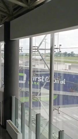 The video shows a view through the glass of what appears to be an airport terminal. Outside, an Ethiopian Airlines airplane is visible on the tarmac. The plane is white with the distinctive Ethiopian Airlines green, yellow, and red stripes on the tail. In the foreground, there's a large sign for "First Citizens Bank" visible through the glass. The glass is supported by metal rods and fixtures. The inside area has modern architecture with visible structural supports. There is also a glass railing along the bottom of the frame. The day is overcast.