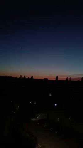 The video shows a darkening sky with a gradient of blues and reds near the horizon, suggesting either sunrise or sunset. Silhouetted against the sky are trees and some indistinct structures. In the foreground, the scene is largely dark, but lights from buildings and streetlights are visible, illuminating parts of a road and parked vehicles. There are no visible people or interactions.