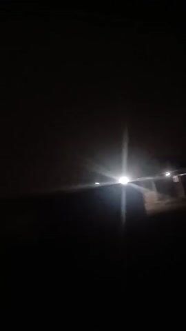 The video is taken at night with very low lighting. A bright light source, likely a street light or vehicle headlights, shines from the upper right of the frame. A dark silhouette of what seems to be a structure or landscape feature occupies the lower portion of the frame. The overall visibility is poor, making it difficult to discern specific details clearly.