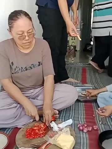 The video shows three people sitting on a mat on the floor, seemingly preparing food. A woman in the foreground, wearing glasses and a brown t-shirt, is chopping red chilies on a wooden cutting board. To her right, a young woman wearing a black hijab and glasses is peeling small red onions. In the background, a person wearing a dark blue shirt and black pants stands, holding a small spoon or utensil and interacting with the woman in the hijab. There are various food items and cooking utensils scattered around them, including a metal tray with small fish, a frying pan, and a container of what appears to be chopped meat. The environment appears to be indoors, possibly a porch or a room with a door and a fan visible in the background.