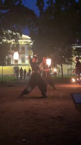 Three individuals are performing a fire-spinning act outdoors at dusk. Two individuals in the foreground are actively spinning fire poi, with flames emanating from the props. They are dressed in dark, loose-fitting clothing. A third individual in the background is also spinning fire poi. Behind the performers, a large, light-colored building with windows is visible, and trees cast shadows on the ground. Several other people are standing in the background, appearing to watch the performance. The ground is covered in dirt or gravel. A dark case or box is visible in the lower right corner of the frame. The sky is a deep blue, indicating twilight.