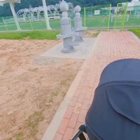 A camera moves forward, showing a person pushing a stroller. The stroller is black and appears to be a double stroller. The camera is mounted on the stroller. In the background, there are two stone fountains with cartoon-like figures on top. The fountains have multiple spouts and basins. Beyond the fountains, there is a grassy area with a fence, a sports field, and hills in the distance. The sky is overcast.