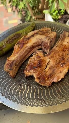 The video shows a close-up shot of grilled meat and pita bread on a grill. The meat appears to be lamb chops, with grill marks visible. A green pepper is also on the grill. The pita bread is golden brown and slightly puffed. The scene is outdoors, with greenery and a tiled patio visible in the background. The video is a short, looped clip with no discernible audio.