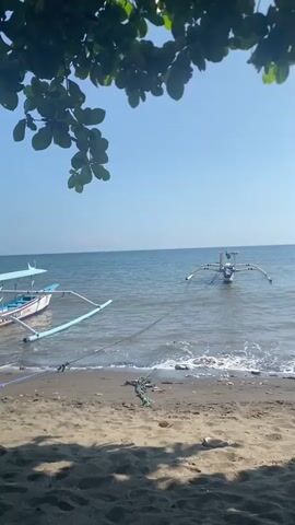 The video shows a beach scene with two boats in the water. The sand on the beach is dark and appears wet in places, with some debris scattered on it. In the foreground, there are shadows cast by leaves and branches from a tree overhead. The ocean is calm and blue, with small waves lapping at the shore. The sky is clear and bright blue. One boat, a traditional outrigger canoe, is anchored closer to the shore. Another similar boat is anchored further out in the water. The video is shot from a low angle, looking out towards the sea.