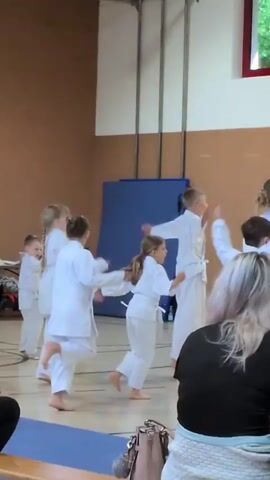 The video shows a group of young children in a gymnasium. They are all wearing white karate uniforms. The children are practicing martial arts movements. Some are jumping, some are punching, and some are kicking. The gymnasium has a wooden floor and brown walls. There is a large blue mat in the background. In the foreground, there are spectators. The video is shaky and the lighting is poor.