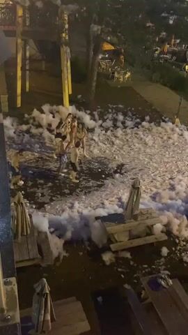 The video shows an outdoor party scene at dusk or night. The ground is covered in a thick layer of white foam, suggesting a foam party event. In the foreground, there are several picnic tables with umbrellas, some of which are closed. A few people are visible near the tables. Further into the scene, a group of people, mostly young adults, are interacting in the foam. They are wearing swimwear and appear to be enjoying themselves. In the background, there are more lights and what appears to be a swimming pool area filled with people. The overall impression is that of a lively, festive atmosphere. There are also some structures visible, possibly part of the venue's infrastructure or entertainment setup. The lighting is dim, with artificial lights illuminating the area.