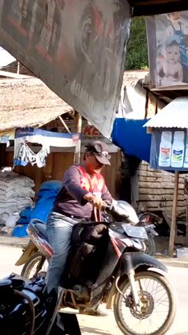 Here's a concise description of the scene:

A man wearing a black cap, sunglasses, a black and red long-sleeved shirt, and jeans is sitting on a black motorcycle parked on a paved street.  He's fiddling with something on the handlebars.  In the background, there's a mix of corrugated metal and thatched roof structures, advertising banners, and a pile of blue tarps. A child walks by.