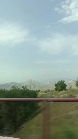 The video shows a view from a vehicle, possibly a car or train, moving through a rural landscape. The camera is positioned at a side window, and the view is slightly blurred due to motion. In the foreground, a red railing or barrier is visible, suggesting the vehicle is on a bridge or elevated structure. Beyond the railing, there are green trees and bushes. In the background, rolling hills and mountains can be seen under a partly cloudy sky. Power lines are visible stretching across the sky. The overall impression is of a scenic drive through a natural environment.