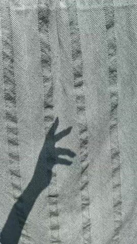 The video shows a shadow of a hand against a textured, striped curtain or fabric hanging outdoors. The hand is in motion, and its fingers are manipulated in various ways, such as forming a fist, pointing, or spreading apart. The fabric appears to be light-colored with darker stripes running vertically. The lighting suggests it is daytime, with sunlight casting the shadow. The background above the fabric is blurry, but hints at greenery and possibly a sky. The bottom of the fabric has a fringe.