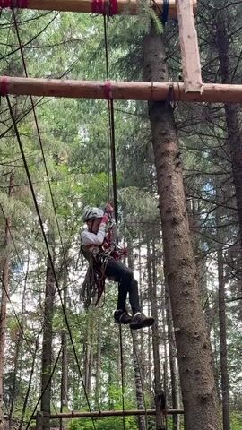 A person wearing a helmet and a harness is suspended in the air from a rope course in a forest. The person is holding onto a rope with their hands and appears to be swinging or moving across the course. The background consists of trees and a wooden structure.