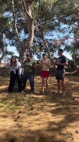 Four people stand in a wooded area on a sunny day.  A woman in sunglasses and a white shirt stands on the left. Next to her is a man in an olive-green shirt. Another man in a white tank top and red shorts stands to his right. The fourth person, a man in a black shirt, stands on the right edge of the frame. They are all talking and gesturing. The ground is covered in dry grass and pinecones. Several trees surround the group. A hammock hangs between two trees in the background.