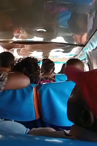 The video shows the interior of a passenger van. Passengers are seated on blue seats with orange trim. A person wearing a red baseball cap is visible in the foreground. The ceiling of the van is reflective, showing distorted images of the passengers. There is some spoken audio.