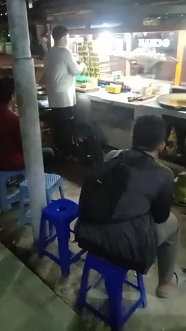 The video shows an evening scene at a street food stall in Kebonsari, Indonesia. Three men are present. One man, wearing a white shirt, stands behind a brightly lit counter, actively preparing food or drinks. On the counter, various items are visible, including stacked containers or boxes, and what appears to be a large frying pan or cooking surface. Two other men are seated on blue plastic stools facing the counter, seemingly customers waiting for their orders. One of the seated men wears a black jacket and has a backpack on. A gas cylinder is visible near the base of the counter. The artificial lighting suggests it is nighttime. In the background, part of a sign with the word "MANDO" can be seen.