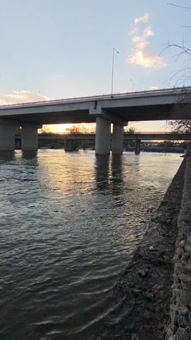 The video shows a fishing lure dangling in front of a river. Initially, the lure is orange and yellow, but it changes to green and yellow. The river is flowing, and there is a bridge in the background. The sun is setting, casting a warm glow on the water. The banks of the river are visible with some vegetation. The sky is blue with some clouds.