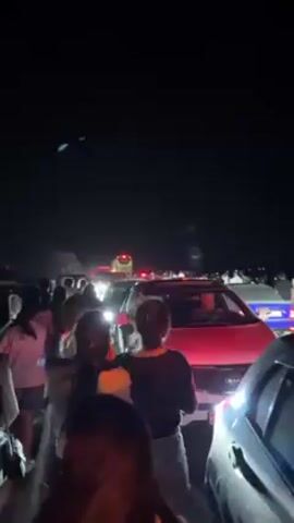 The video shows a large group of people standing on what appears to be a road or highway at night. Cars are present and stopped in both directions. Some cars have their brake lights on. Many people are facing towards an unknown point in the distance, possibly looking at an event or spectacle. It is difficult to determine the exact environment due to darkness, but it seems to be an open area with a gathering of people and vehicles.
