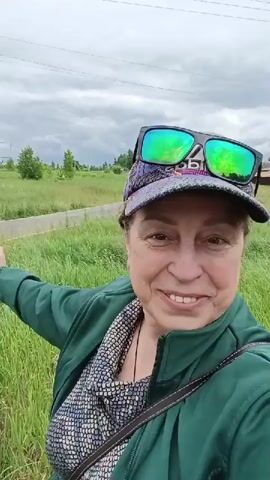 A woman in her late 40s or early 50s, wearing a green jacket, a patterned shirt, a cap with sunglasses perched on top, and a bag, smiles at the camera. She is standing in a grassy field with a road in the background. Power lines and trees are visible in the distance. The sky is overcast, suggesting a cloudy day. The woman is pointing with her left arm, indicating something out of frame. Her expression is cheerful and engaging. The setting appears to be a rural or semi-rural area in Грибчиха, Russia. The time of day is difficult to determine precisely due to the overcast sky, but it seems to be daytime. The overall mood is positive and inviting.