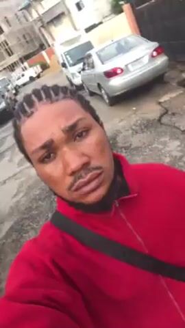 A man takes a video of himself while standing on a street. He has cornrow braids and a small beard. He is wearing a red zip-up jacket and a black crossbody bag. He makes a hand gesture towards the camera. A silver car is parked behind him. There are buildings and trash cans visible in the background. The street appears to be in disrepair.