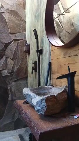 The video shows a modern bathroom. The walls of the shower are made of large, irregularly shaped rocks, creating a rugged, natural aesthetic. A black shower head and faucet are mounted on the wall. A stone sink sits on a wooden countertop. A round mirror is mounted above the sink. A soap dispenser is also attached to the wall. The shower area is illuminated by a skylight above. There are no people visible in the video. The camera moves around the room to capture different angles of the bathroom.