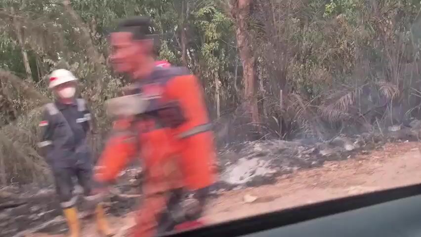 The video shows a view from inside a vehicle. In the foreground, the side of the vehicle's interior is visible, including a seatbelt and a plastic bottle of water on a shelf. Outside the vehicle, the scenery is blurry and suggests a dusty or smoky environment. Trees and vegetation line the side of a dirt road. In the distance, several people are gathered on the side of the road, some standing and some on motorcycles. The overall scene appears to be in a rural or undeveloped area. There is evidence of smoke or dust in the air, obscuring some of the details of the environment and the people.