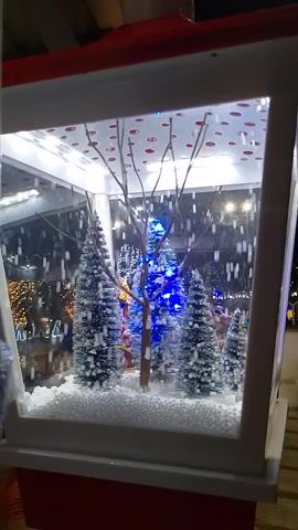 The video shows a Christmas display inside of a white box with a clear front. The display features fake snow, small evergreen trees lit with various colored lights, a figure of Santa Claus, and a bare tree branch. Simulated snow falls within the box. The backdrop of the inside of the box is white with red polka dots. The overall scene evokes a Christmas or winter theme.