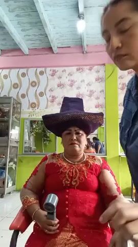 A woman wearing a large, flat-brimmed purple hat and a red sequined dress sits in a chair. Her left hand holds a black device. Another person, partially visible from the shoulder up on the right, adjusts the hat on the seated woman's head. The background appears to be an indoor setting, possibly a salon, with wallpaper featuring a floral pattern. A ceiling with exposed beams is visible above.