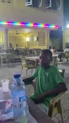 The video shows a brightly lit outdoor dining area, possibly a restaurant or bar, at night. Several people are seated at tables, and a building with a yellow facade and a colorful string of lights is visible in the background. In the foreground, there are several tables with plastic bowls and bottles of water and other beverages. Two young individuals are visible at one of the tables; one is wearing a green shirt and sitting idly, while the other wears a blue patterned shirt and appears to be eating. Other patrons are scattered throughout the dining area, some engaged in conversation. The scene suggests a casual, social gathering in an outdoor setting.