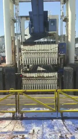 A ferry ramp is in the center of the frame, with a large blue crane mechanism above it. The ramp has a series of metal prongs that are extended. The scene is likely a ferry terminal, with the ocean or water visible in the background, along with some buildings and possibly cliffs. There are some people visible in the distance to the left of the frame, and some heavy black fenders on either side of the ramp. A yellow metal railing is in the foreground, obscuring part of the view. The sky is blue and clear, suggesting it is daytime. The overall impression is of a functional, industrial port environment.