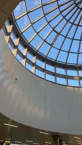 This media file is a video looking up through a glass-domed ceiling towards a bright blue sky. A tall, modern, yellow-toned apartment building is visible through the transparent panels of the dome. The building has many windows and appears to be in an urban setting. The sunlight casts shadows across the interior walls of the space beneath the dome, indicating it is daytime. The lighting suggests it could be late afternoon or early morning, with the sun at an angle. The interior appears to be a large, open space, possibly a shopping mall, atrium, or public building, with white, curved walls and some overhead lighting fixtures visible at the bottom of the frame. There is no visible text in the video. The overall impression is one of modern architecture and a clear, sunny day.