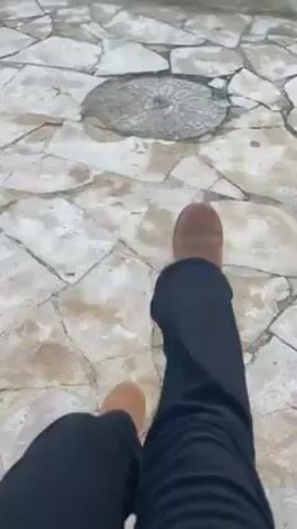 The video begins with a ground-level shot of a person's foot resting on a stone-tiled surface. The camera then pans upward to reveal a park landscape. There is a small body of water in the mid-ground, surrounded by grass and trees. Buildings are visible in the distance under a pale blue sky with some clouds. The camera pans to the right, showing a grassy hill with stairs, a single tree, and palm trees in the background. A black dog with white markings on its legs walks across the stone tiles toward the camera. The scene is bathed in soft, natural light, suggesting early morning or late afternoon.