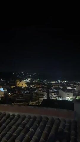 A nighttime aerial view captures a coastal town illuminated by numerous lights. The camera pans across a beach and the adjacent sea, where reflections of the lights shimmer on the water's surface. Buildings and lights are visible along the coastline, and on hillsides in the distance. In the foreground, a dark rooftop made of terracotta tiles is partially visible.