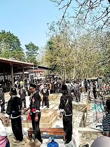 A group of people, many wearing traditional clothing, gather outdoors around a large white conical object. Some individuals are dressed in black martial arts uniforms with blue sashes. In the background, there is a building with a tiled roof and trees. One person in the foreground is holding a pineapple and other decorative items. The atmosphere appears to be celebratory or ceremonial.