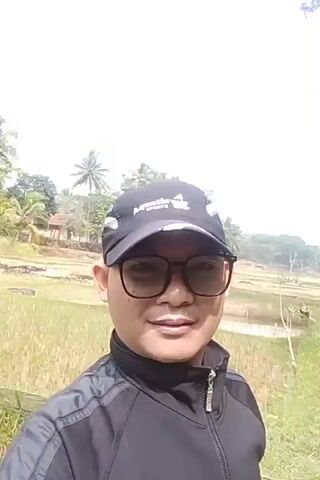A man wearing a black baseball cap and black-rimmed glasses smiles at the camera. He is wearing a black zip-up jacket with gray stripes on the sleeves. Behind him, a field of green vegetation, possibly rice paddies, stretches into the distance. There are trees and a house in the background. The sky is bright and overcast. The video appears to be taken outdoors during the day.