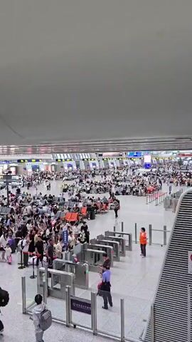 The video pans across a crowded indoor space, likely a train station or airport terminal. Numerous people are visible, many of them seated on rows of benches, while others stand in queues or walk through the area. Several ticket gates are present, with some people passing through them. In the background, digital display boards show information, and the architecture of the building features a high, modern ceiling with prominent lighting fixtures. The overall atmosphere appears to be one of busy travel and transit.