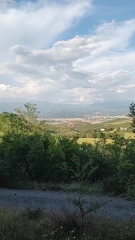 The video opens with a panoramic shot from a hillside, looking out over a valley. In the foreground, green trees and bushes are visible, with a dirt path at the very bottom of the frame. In the middle distance, a town or city with numerous buildings spreads across the valley floor, nestled between rolling hills and distant mountains. The sky above is partly cloudy.

The camera then slowly zooms in on the scene. As it tightens its focus, a prominent, large white cross becomes clearly visible on a distant, tree-covered hillside, overlooking the town from above. The zoom continues, making the cross the central subject of the frame. The overall scene depicts a peaceful, mountainous landscape with an urban settlement and a significant religious symbol on a hilltop, indicative of the location in Selishte, Bulgaria. The lighting suggests daytime, possibly late afternoon, with soft, natural light. No text is visible in the video.