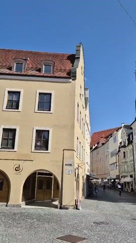 The video shows a street scene in a European city. On the left, a light yellow building with a red tiled roof and arched doorways is visible. In the distance, a tall, narrow, brick clock tower with a spire stands against a clear blue sky. To the right of the tower, several multi-story buildings with ornate facades line the street, featuring rows of windows and decorative architectural elements. The street itself is paved with cobblestones. Several people are walking on the street. Some are dressed in casual clothing, while others appear to be wearing more traditional garments, possibly indicating a cultural event or festival. Two women in long dresses are walking towards the viewer, and a group of people are walking away from the viewer further down the street. The scene is bathed in sunlight, suggesting it is daytime.