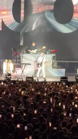 This video shows a concert performance. A band is on stage, with one person in the foreground playing a guitar and singing, dressed in white. There are other band members behind him, and the stage is lit with bright lights and smoke. The audience is visible in the foreground, with many people holding up their phones to record the performance. The overall environment appears to be an outdoor concert venue at night. There are signs visible at the top of the stage that read "FLITZ COLLECTIVE" and "MOBY LIVE".