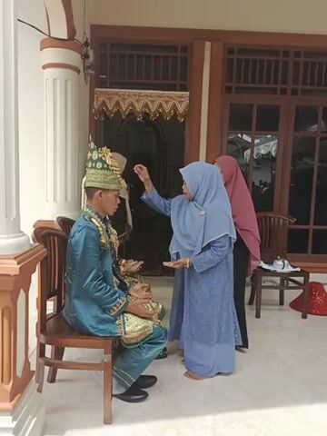 A groom wearing an ornate turquoise outfit and headdress sits in a chair outdoors. Three women in hijabs and long dresses stand around him. The woman nearest to him is reaching towards his head. They are positioned in front of a house entrance decorated with gold and white ornaments. The shot is slightly tilted.