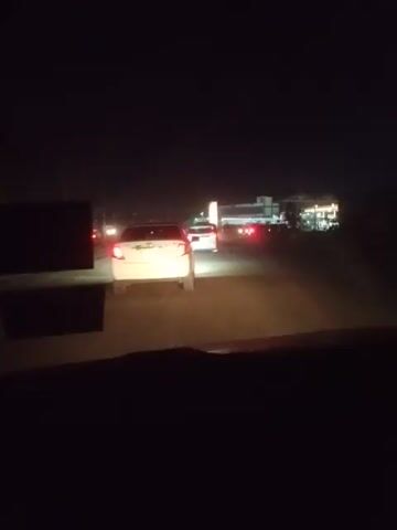 A video shows a road at night. The camera is likely inside a vehicle, focused on traffic ahead. There are several vehicles on the road, including a white ambulance with its lights flashing. A building with bright lights is visible in the distance. The environment appears dark and the road is illuminated by vehicle lights.