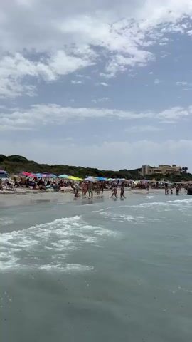 The video shows a crowded beach with many people sunbathing and swimming. The water is clear and blue, with waves crashing on the shore. There are umbrellas set up on the sand, providing shade for beachgoers. In the background, there are buildings and trees. The sky is partly cloudy, with some scattered clouds. Several boats are visible in the distance. People are walking along the shoreline, and some are playing in the water. The overall scene depicts a lively and enjoyable day at the beach.